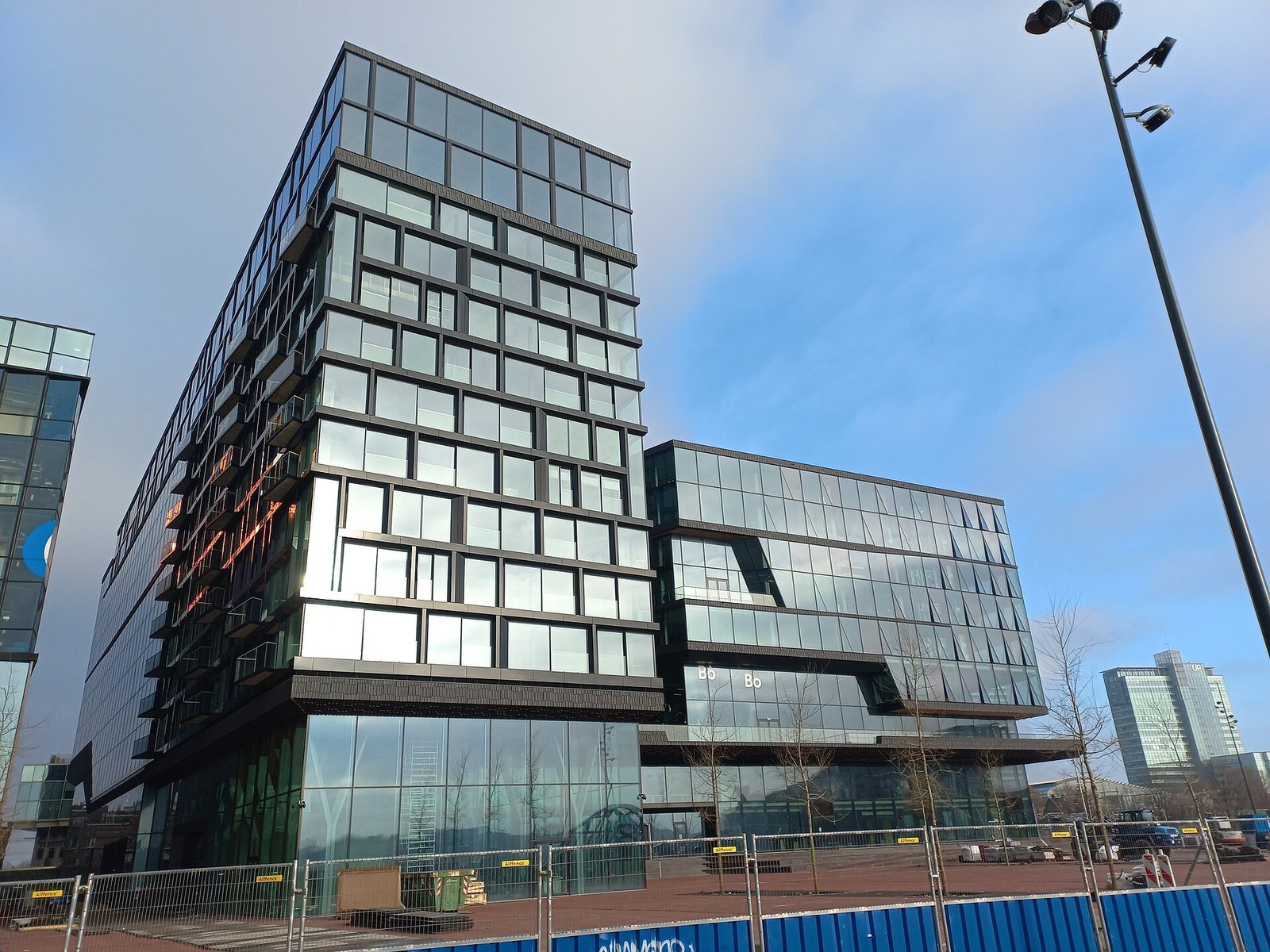 Booking.com's headquarters on the Oosterdokseiland in Amsterdam: two glass office blocks with dark window frames under a blue sky, with construction fencing at street level.