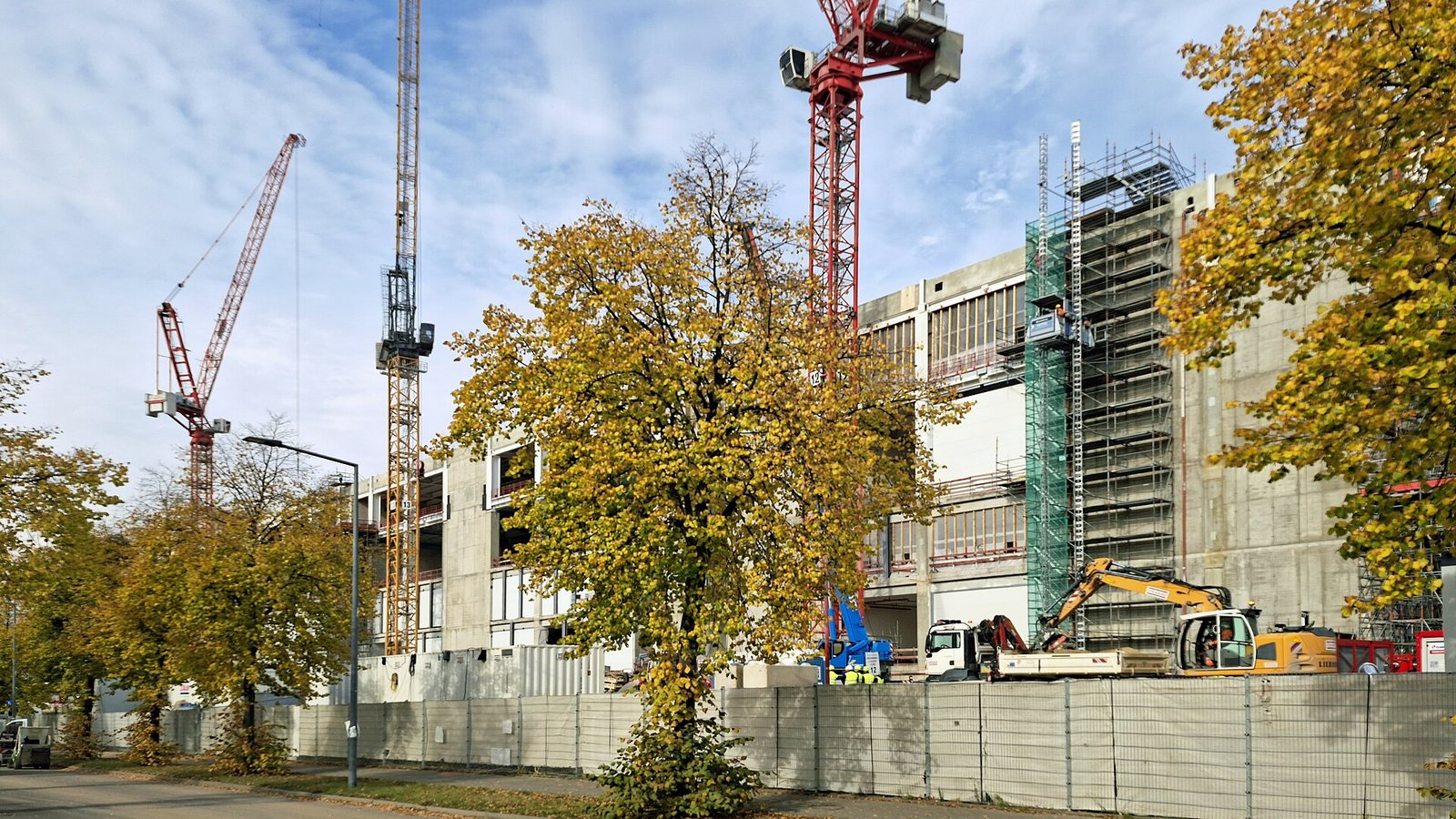 Construction site of the ESMC chip factory in Dresden: grey concrete factory buildings with scaffolding, three tower cranes, an excavator and a lorry behind a site fence, with autumn trees in the foreground.